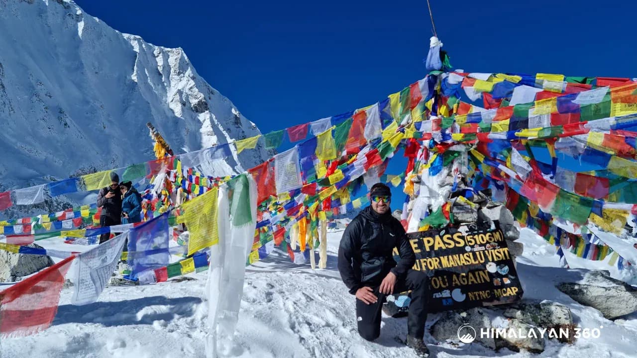 A trekker beside the Larkya La (5,106M) sign under prayer flags and snow on the Manaslu Circuit